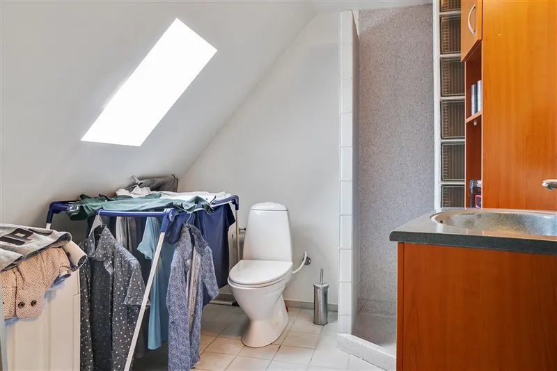 Lyst og rent badeværelse med toilet, vask og brusekabine samt tøjstativ og loftsvindue