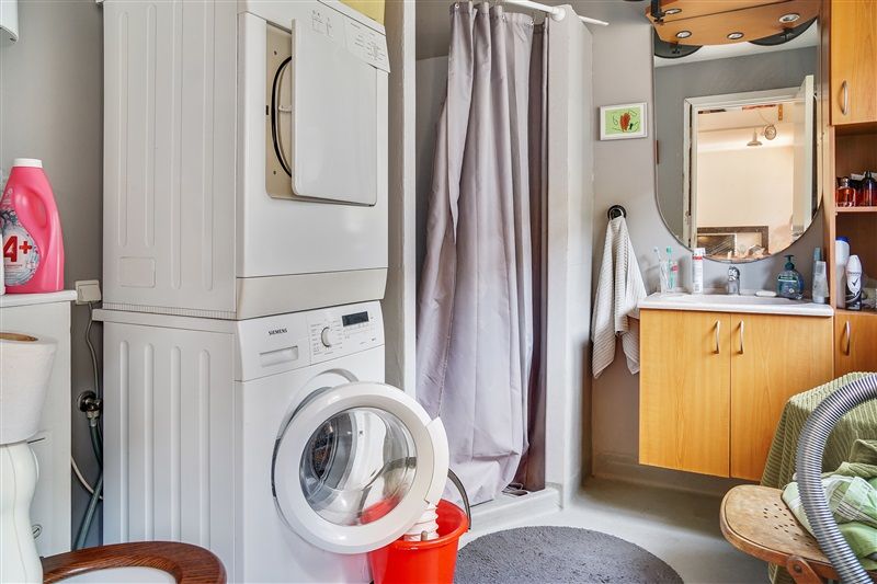 Vaskerum med hvid vaskemaskine og tørretumbler samt toilet, håndklæde, rengøringsmidler og en spejl i et lille badeværelse