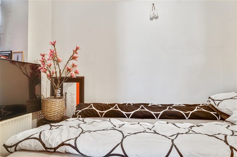 Et soveværelse med en tv-skærm, en vase med lyse pink blomster, et tekstilbillede og en sengeliggende.