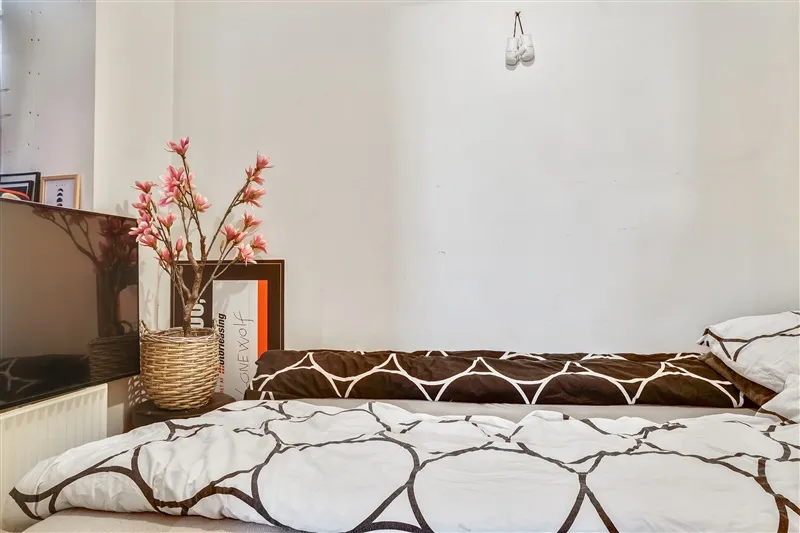 Et soveværelse med en tv-skærm, en vase med lyse pink blomster, et tekstilbillede og en sengeliggende.