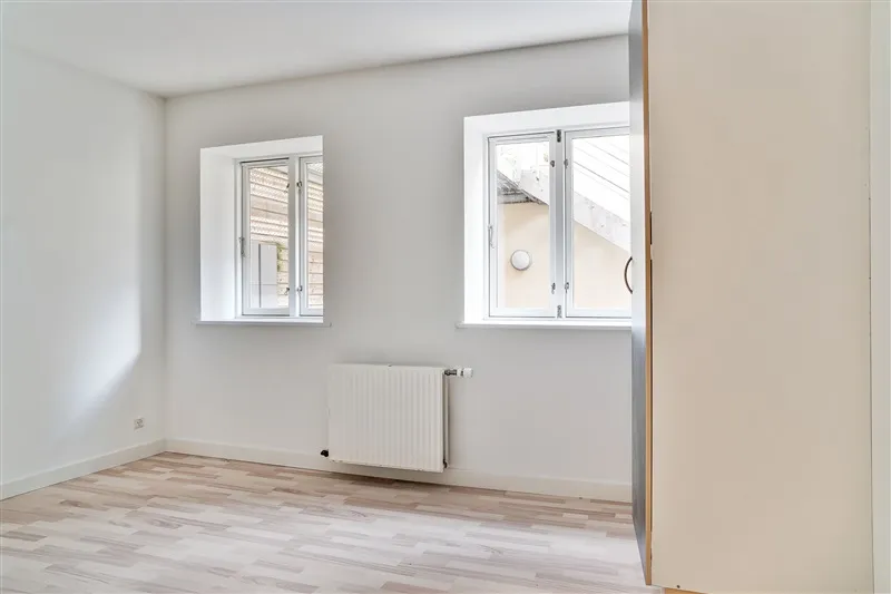 Et tomt rum med hvide vægge og loft, to vinduer og en radiator samt en åben dør