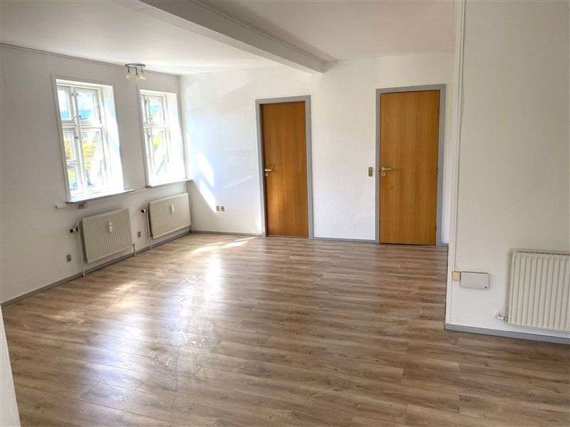 Værelse med to døre, hvide vægge, lyse trægulve og to vinduer med lysegardiner. To radiatorer og to loftslamper i værelset