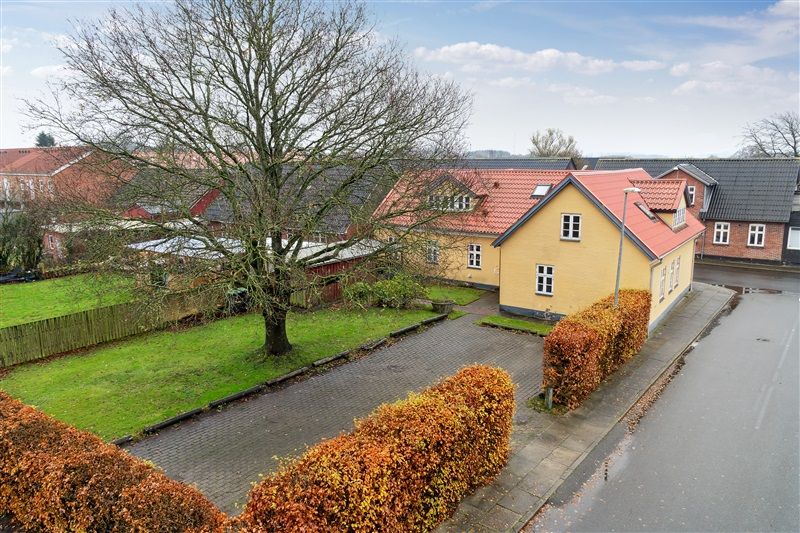 En stor gul villa med rød tagdækning og en lille indkørselsvej. En stor trækrone med gule og orange blade. Et grønt græsfelt