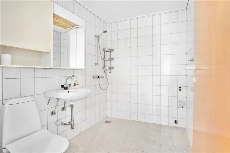 Hvidt badeværelse med toilet, hvidt wask og håndklædeholder samt en hånddusch med en håndklædestol.