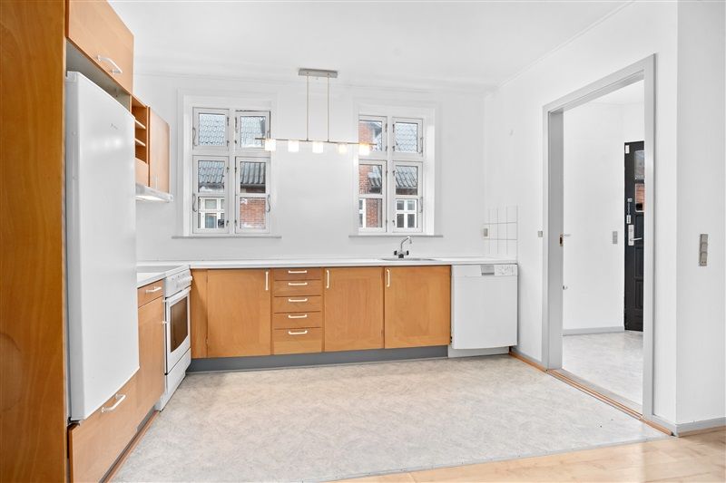 Et køkken med hvide vægge, træskabe, hvidt køleskab, lysekroner, spisestue, og moderne indretning