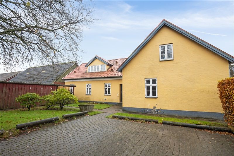Gul rækkehus med tegltag og terrasse står i en fredelig gade i Skovlund med grønne planter og træer
