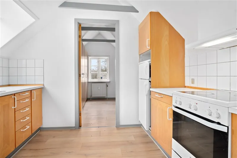 En køkken med hvide vægge og træfronter, et køleskab og en komfur samt en vinduesåbning i baggrunden