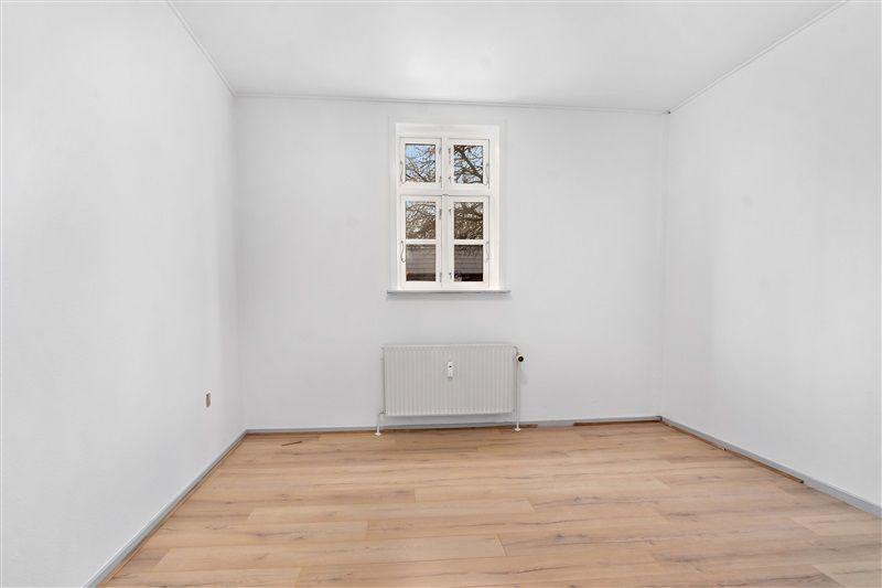 Lyst og rummeligt værelse med hvidt loft og hvide vægge samt gult trægulv. Værelset er fuldt udstyret med radiator og vindue