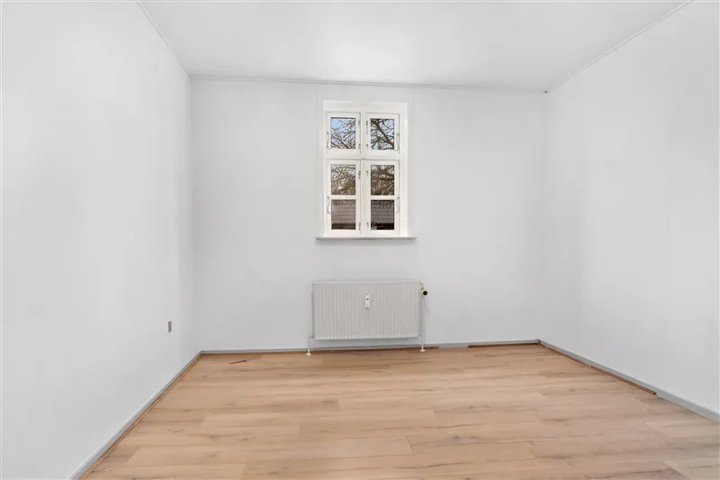 Lyst og rummeligt værelse med hvidt loft og hvide vægge samt gult trægulv. Værelset er fuldt udstyret med radiator og vindue