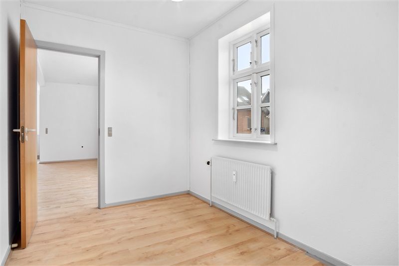 Et rensvejset og moderne værelse med hvide vægge og trægulv, samt et vindue og en radiator langs væggen