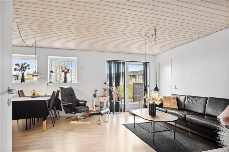 Et moderne og rummeligt stueområde med sort lædersofa, højloft, lyse trægulve og lysegardiner