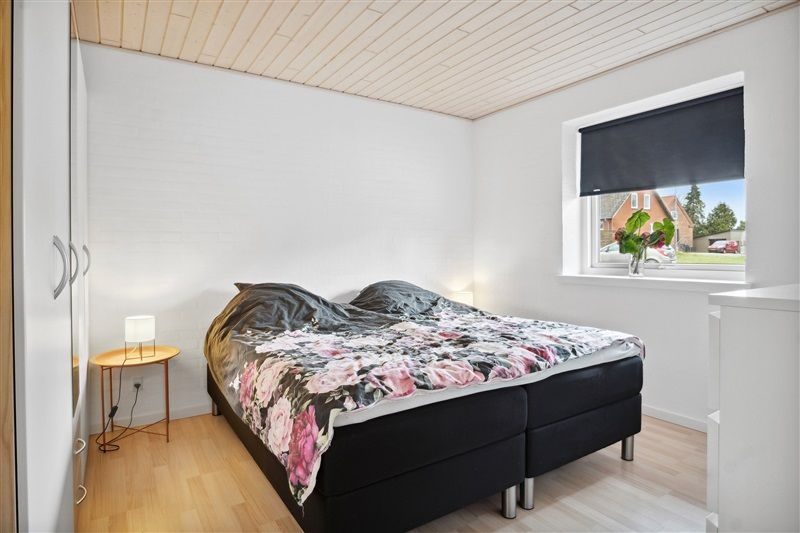 Soveværelse med trægulv og hvide vægge, dobbeltseng med blomstret dynebetræk, bord med lampe og vase med røde blomster samt