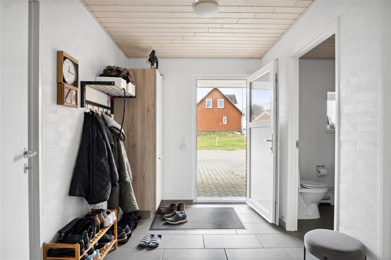 Indtrædensområde med åben dør, toilet, køkken og garderobe med hængende tøj og sko samt klokker og plante i hjørnet