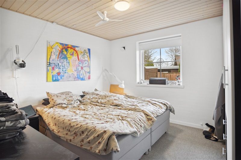 Soveværelse med dobbeltseng og hvidt loft og vægge. Sengetøj med mønstre og hvidt sengehoved. Maleri på væggen og klædeskab