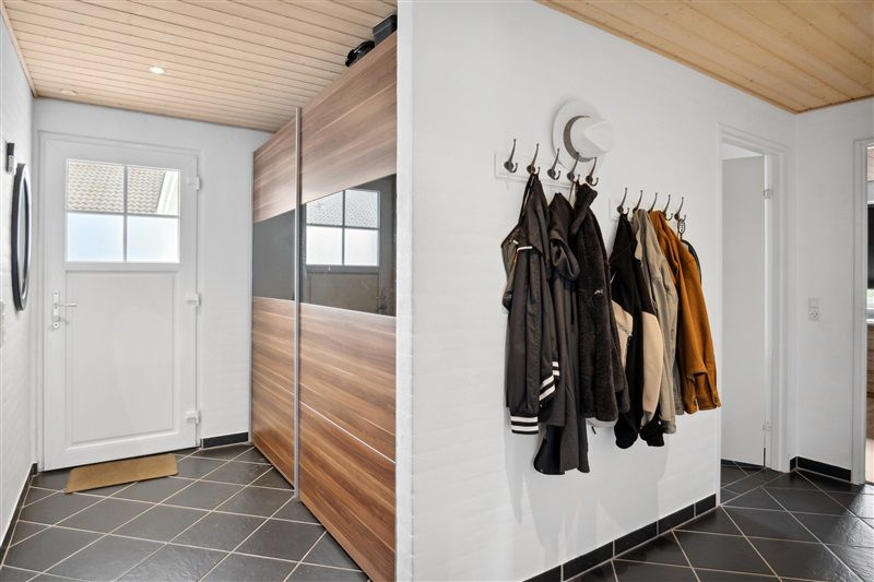 Moderne entre med hvide vægge og træloft. Sort-hvidt gulv med tæppe ved indgangen. Hængeklæder og hatte på væggen med lyse