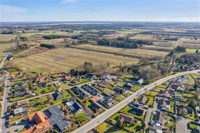 Landsby med rækkehuse, gadekøkken, grønne marker og skove i baggrunden. Vej og biler, fredelig atmosfære og blå himmel.