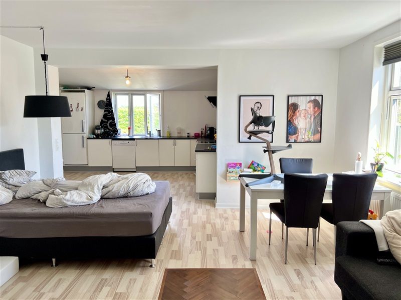 Moderne lejlighed med åbent køkken, trægulv og lyse lofter. Billeder hænger på væggen og en sofa med pude ligger mod vinduet