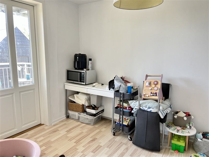 Et værelse med et hvidt køkkenbord og en mikroovn ovenpå. Væggene er hvide, og der er et trægulv. Der er en stol med høj