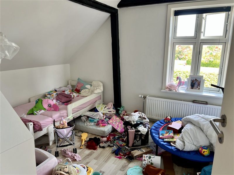 Større børneværelse med to senge, et legeområde og en åben dør til et opholdsrum med radiator og vindue med udsigt til