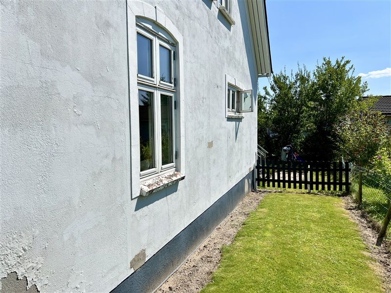 Hvid malet hus med tre vinduer og skrånende tag, liggende i et område med græs og træer