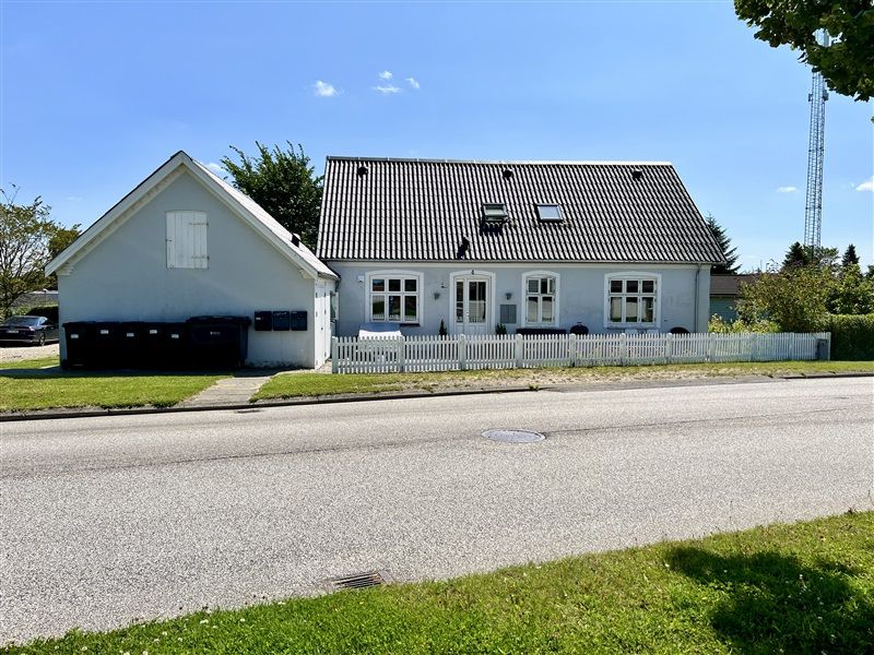 To hvide hus med skrånende tag, foran en hvid skur, udstyret med et lille skur og skraldespande langs gaden med en grøn mark