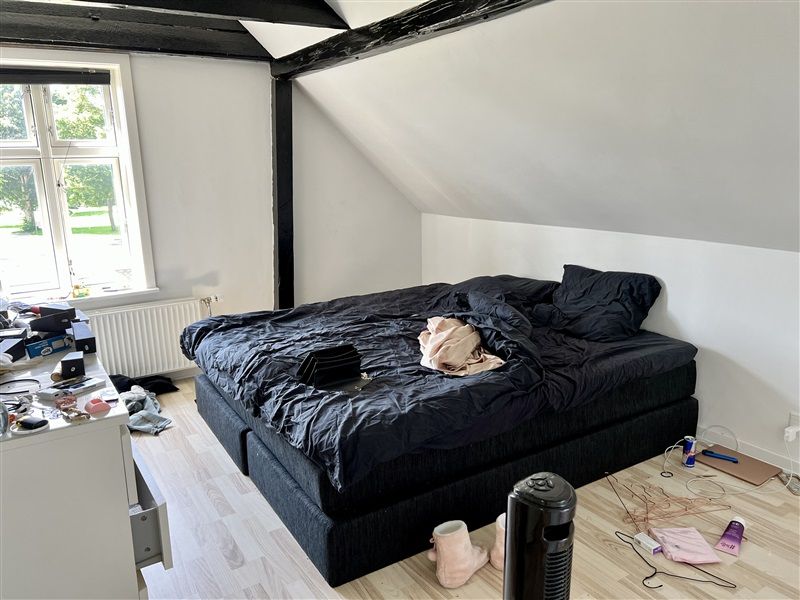 Et ubeboet værelse med sort sengebetræk og tøj på gulvet og et snavset skrivebord med diverse genstande