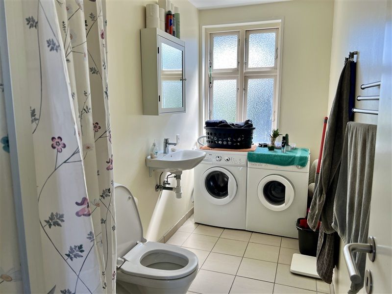 Toilet med brusebad og 2 hvide tøjvaskere samt vask med 2 håndklæder og rengøringsprodukter