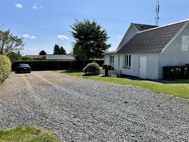 Hus med hvide vægge og sort tag. Større terrasse og skovløb til parkering. Skov og hæk adskiller vej fra gårdsplads.