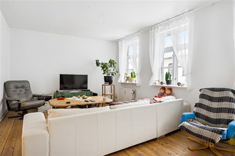 Lejlighed med højloft og lysevinduer, hvor der er en hvid sofa, trægulv og en skuffe i væggen