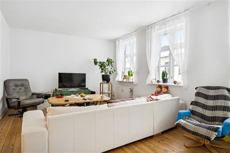 Lejlighed med højloft og lysevinduer, hvor der er en hvid sofa, trægulv og en skuffe i væggen