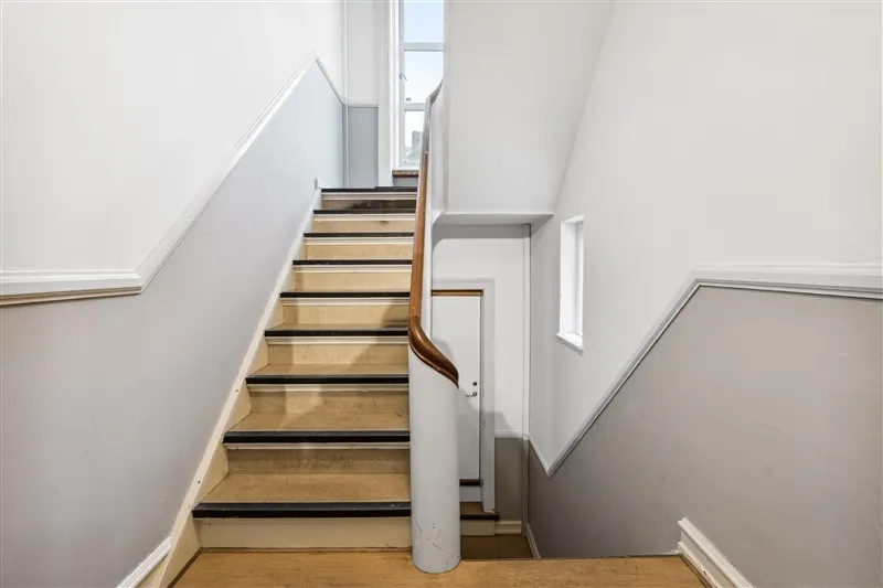 En trætrappe med lysegrå vægge og hvide stiger. Træstigerne er lysebrune og fører op til første sal.