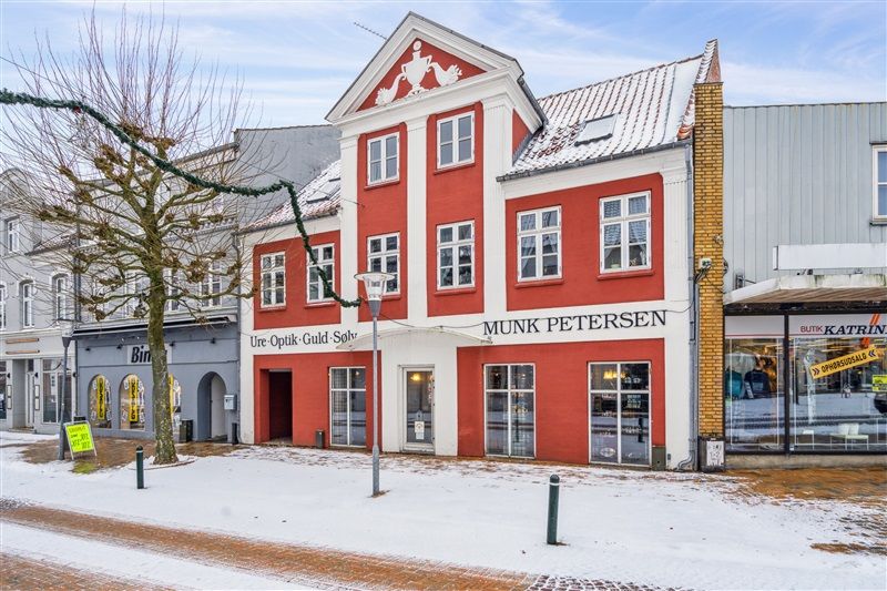 Butik Munk Petersen er i rødt og hvidt og har mange vinduer og en stor skilt, der viser navnet. Sne dækker jorden, og der er