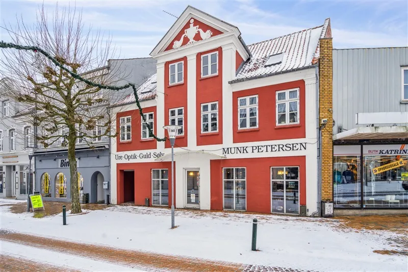 Butik Munk Petersen er i rødt og hvidt og har mange vinduer og en stor skilt, der viser navnet. Sne dækker jorden, og der er