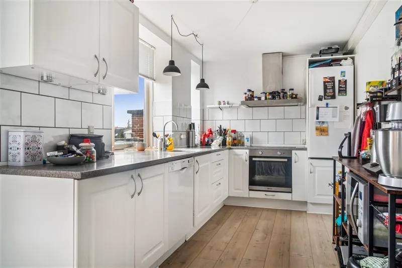 Rent og moderne køkken med hvidt køkkeninventar, hårdt trægulv og køkkenmøbler med håndtag.