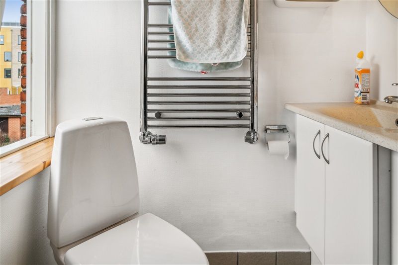 Hvidt toilet i moderne badeværelse med vindue, håndklædeholder og håndvask med rengøringsmidler