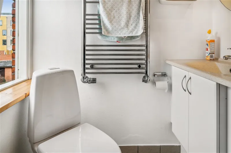 Hvidt toilet i moderne badeværelse med vindue, håndklædeholder og håndvask med rengøringsmidler