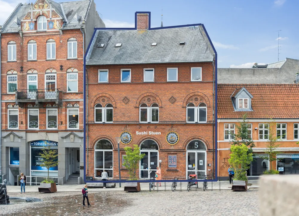 Sushi Stuen er placeret i et bygningskompleks i Svendborg, med en stor glasdør og vinduer, flere planter og cykler parkeret