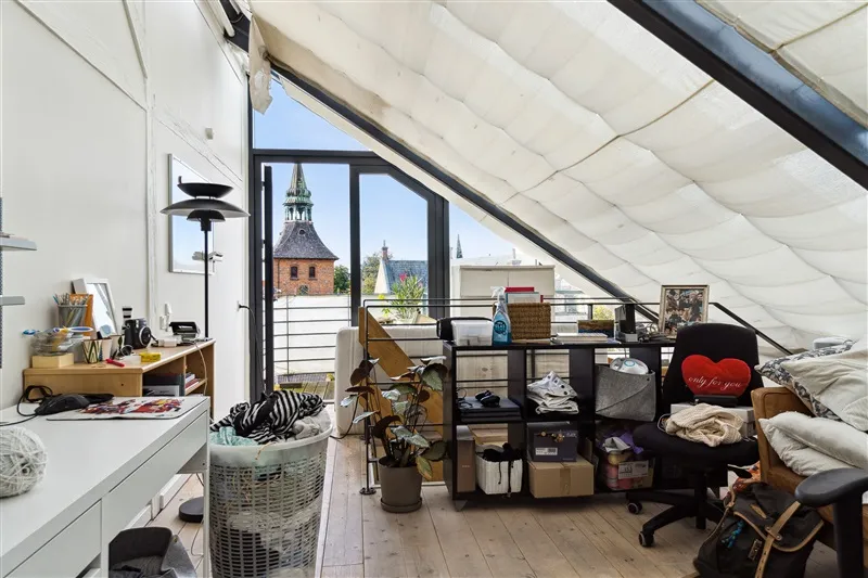 Rommelig stue med loftsrum og udsigt til gadebaner, lyse hvide vægge, trægulve, lysekroner, plante i potte og træhylder med