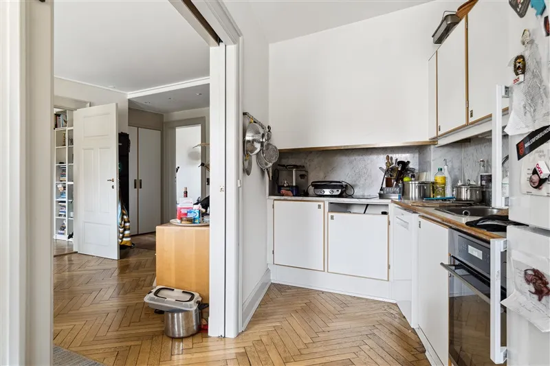 Køkken med hvide vægge og trægulv, åbne skabe og komfur, køkkenredskaber, ølflasker og lysekroner, køkkenbord med flasker og