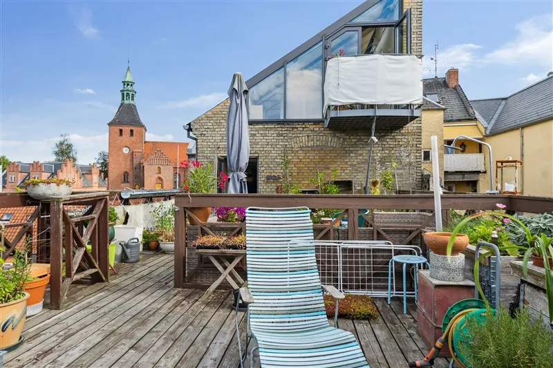 Foto af en balkon med udsigt til Svendborgs Torvet og kirke. På balkonen er der en stuesofa, et bord og flere potter med