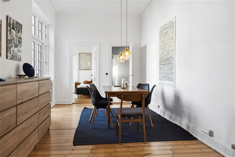 Et lyse og minimalistisk indrettet spisestu med højt til loftet og lyse gulvbelægning. Bord med fire stole og to lysekroner.