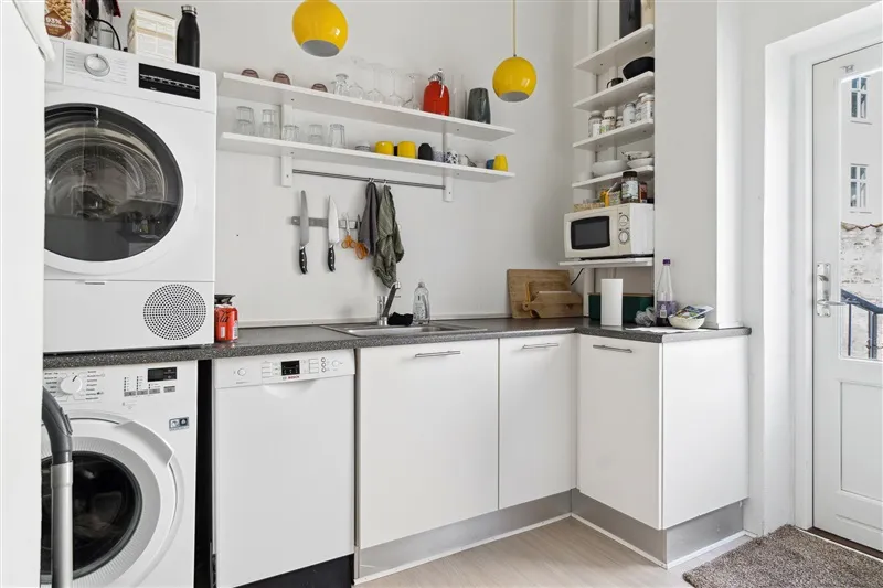 På dette billede ses et rent og velholdt køkken med hvide møbler, to vaskemaskiner og en mikrobølgeovn samt en åben dør til