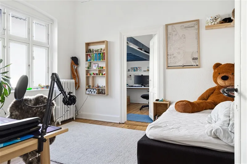 Et soveværelse med hvidt vægmaleri og trægulv, hvor der er en dobbeltseng med hvid dyne og et stort teddybjørn i samme farve