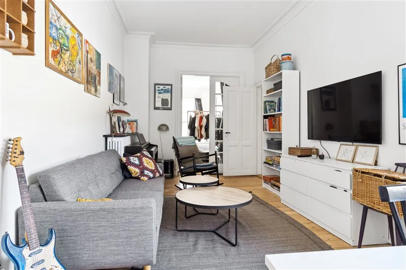 Et moderne stue med lyse vægge, en grå sofa, et gult tæppe, en hvid væskab og en stor tv-skærm