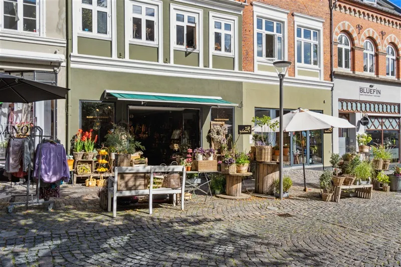 Blomster og tøj sælges i en butik i Svendborg, med et skakbrætsmønsteret gulv og en paraply og stole udenfor