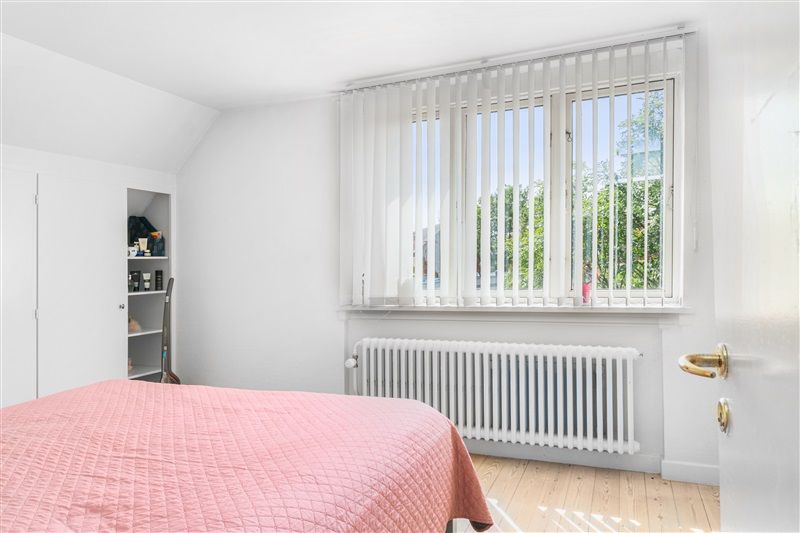 Soveværelse med lyse trægulve, hvidt sengetæppe og pink pudebetræk, hvidt radiator, lysgrå gardiner og højt vindue med