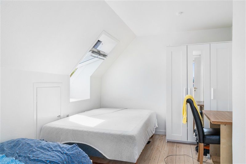 Et soveværelse med hvidt loft og vægge, et dobbeltseng og et skrivebord med en stol og en garderobe med spejl.