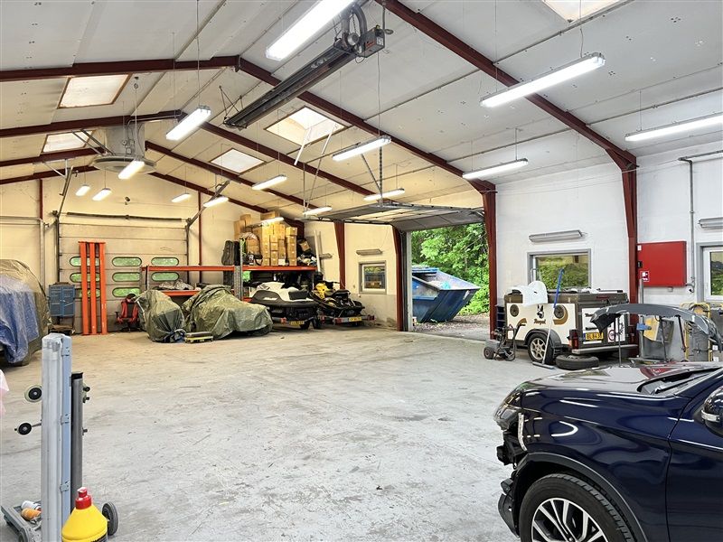 Store garage med biler, ATV, skovmaskiner og værktøj under forskellige loftsmonterede lysarmaturer