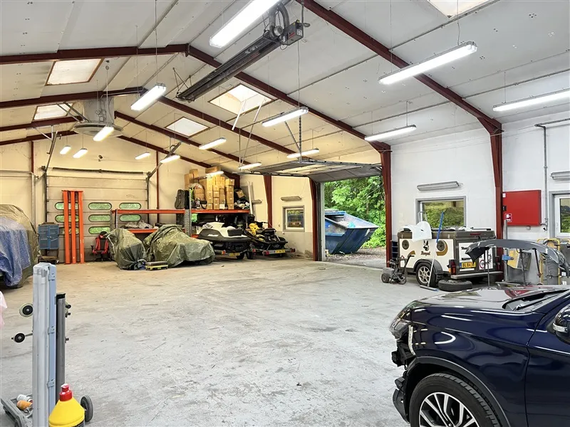 Store garage med biler, ATV, skovmaskiner og værktøj under forskellige loftsmonterede lysarmaturer