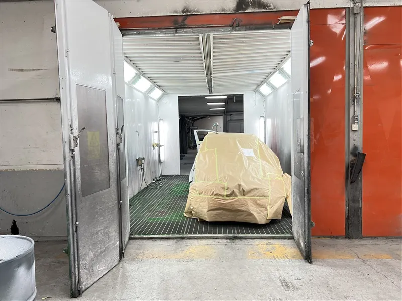 En bil i en malingskammer med hvide vægge og rød dør er dækket med en brun plastpose med gule og grønne linjer